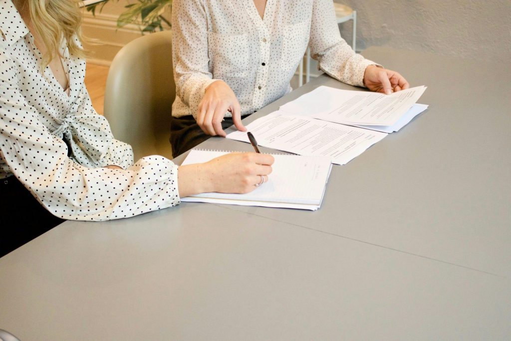 mujer firmando en una hoja de papel blanco de impresora junto a otra mujer a punto de tocar los documentos